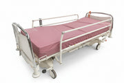 A white hospital-style adjustable bed with a pink mattress, shown indoors against a blue patterned backdrop and concrete floor.