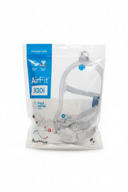 A ResMed AirFit  X30i CPAP mask in its original retail packaging, featuring a clear plastic face mask with a white strap and tubing, sealed in a white and blue bag on a dark gray carpeted floor.