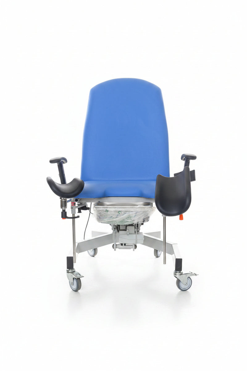 A medical examination table with a blue padded backrest, black armrests, and a wheeled base, photographed in a warehouse or storage area with boxes and equipment around it.