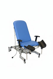 A medical exam table with a blue padded seat and backrest on a white metal frame, photographed in a warehouse or clinic setting with other equipment and cardboard boxes visible around it.