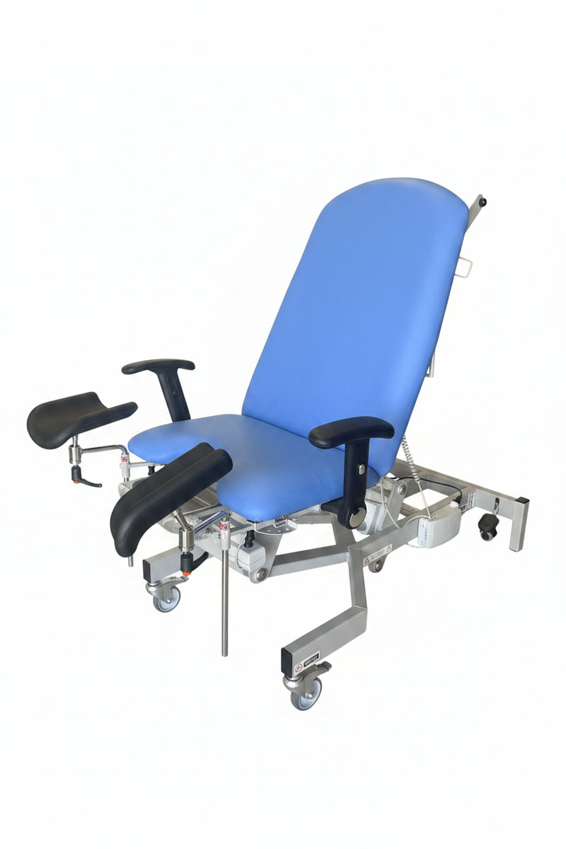 A medical-style reclining examination chair with a bright blue padded seat and back, black armrests, and a white metal frame on casters, photographed in a warehouse or storage area with boxes and other equipment around it.