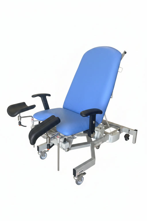 A medical-style reclining examination chair with a bright blue padded seat and back, black armrests, and a white metal frame on casters, photographed in a warehouse or storage area with boxes and other equipment around it.