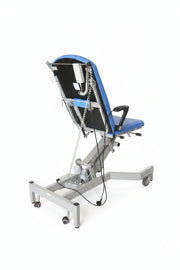A medical-style hospital bed or tilt table with a black and blue backrest, mounted on a white metal frame with casters, photographed in a warehouse or storage area with boxes and equipment in the background.