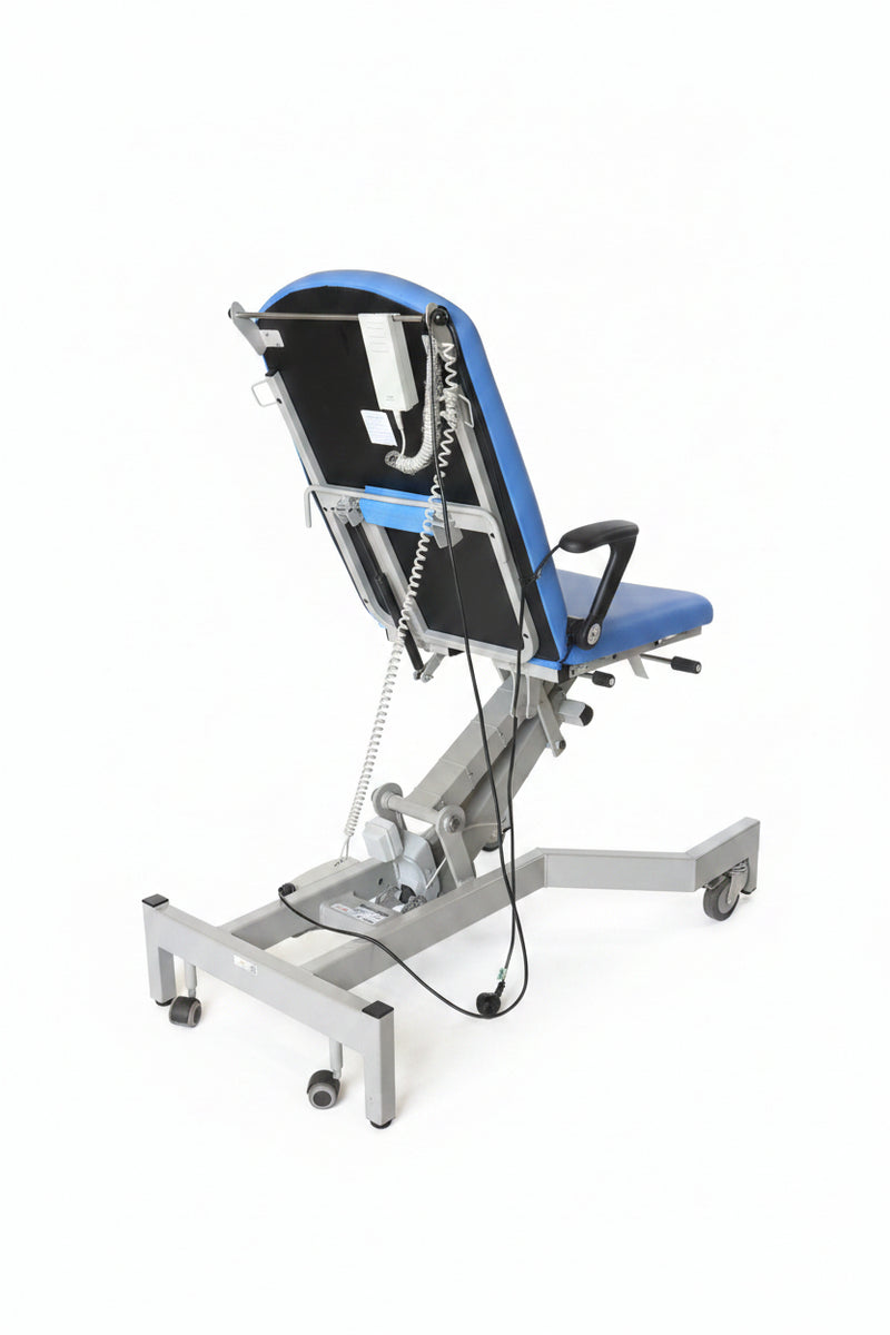 A medical-style hospital bed or tilt table with a black and blue backrest, mounted on a white metal frame with casters, photographed in a warehouse or storage area with boxes and equipment in the background.